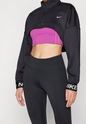 Femme portant une veste courte Nike noire, un soutien-gorge de sport violet et un legging noir taille haute sur un fond uni.