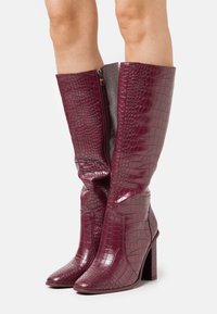 Bottes bordeaux montant jusqu'au genou en matériau verni à motif croco embossé, avec un bout pointu et un talon bloc. Dotées d'une fermeture éclair latérale pour un port facile.