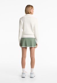 Witte gebreide trui met een geribde zoom, gecombineerd met een groene skort met witte bies. Model draagt witte sneakers en sokken.
