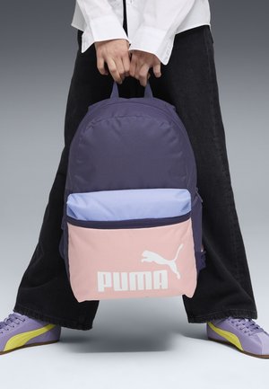 Person holding a purple, light blue, and pink Puma backpack, wearing black pants and purple sneakers with yellow stripes, standing on gray floor.