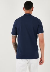 Marinsko modra polo majica s kratkimi rokavi, obrobljena s svetlo modro, z ribasto ovratnikom. Gladek material z oprijetim krojem.