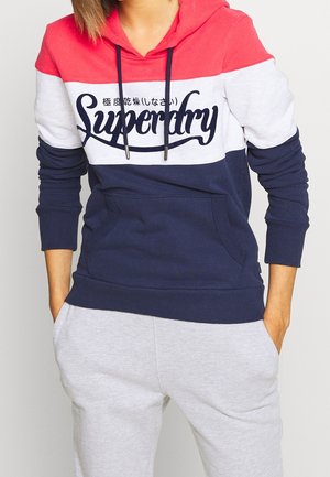 Person wearing a color-blocked hoodie in red, white, and navy with "Superdry" text, paired with light grey sweatpants and hands in pockets.
