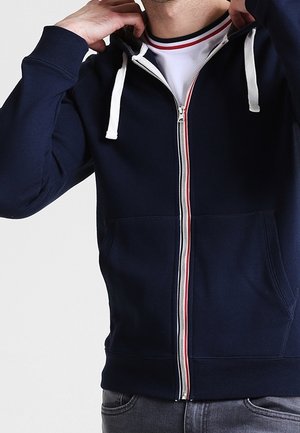 Mężczyzna poprawiający kaptur granatowej bluzy na zamek z białymi sznurkami, w białym T-shircie z czerwono-czarnym wykończeniem kołnierza.