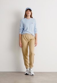 Kvinna står klädd i en ljusblå keps, ljusblå långärmad topp, beige joggerbyxor och vita sneakers mot en vit vägg.