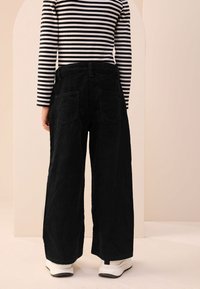 Schwarze Cordstoff-Hose mit weitem Bein und zwei Gesäßtaschen, kombiniert mit einem schwarz-weiß gestreiften Langarmshirt und weißen Turnschuhen.