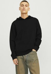 Hoodie noir à coupe décontractée, avec capuche à cordon de serrage, poignets côtelés et un petit patch logo sur la manche gauche. Porté avec un jean clair.
