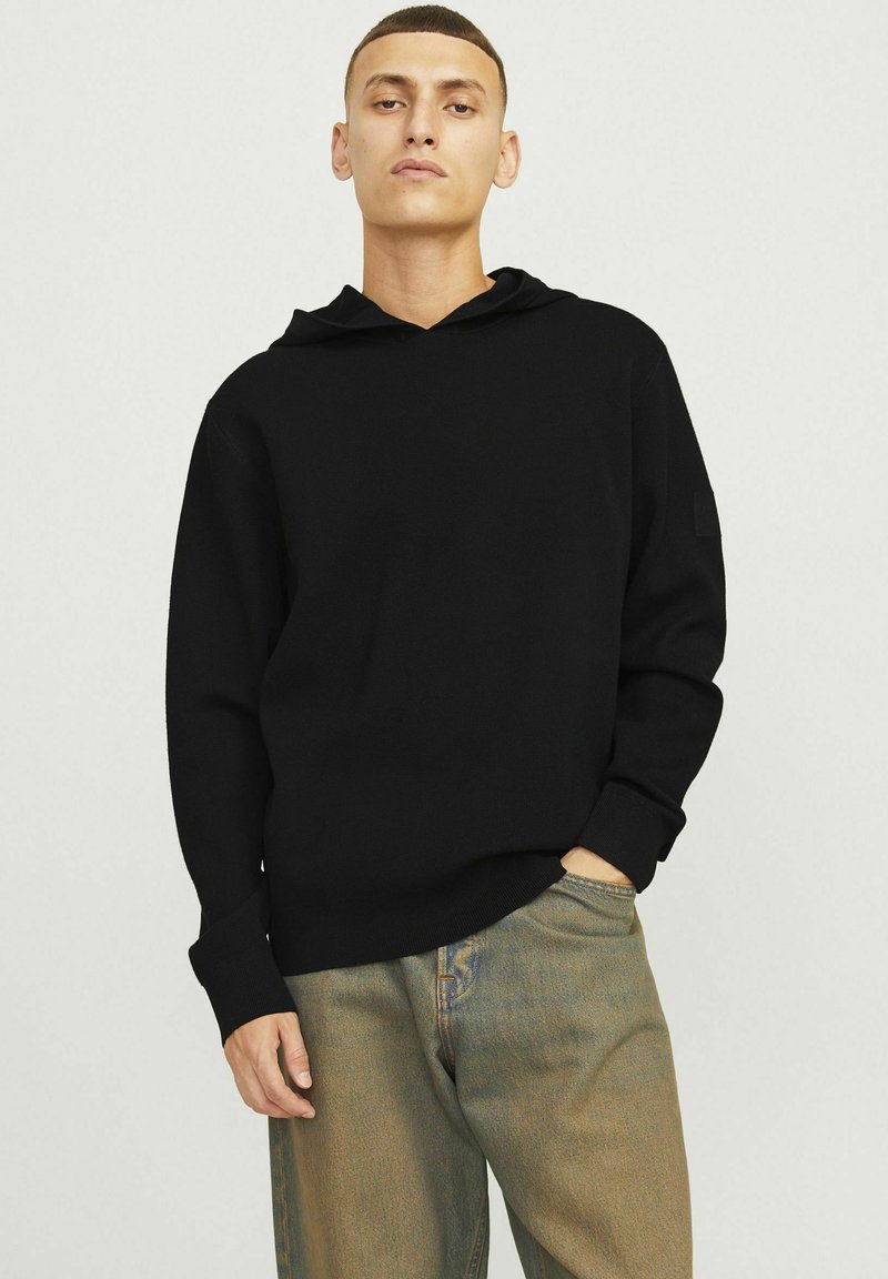 Hoodie noir à coupe décontractée, avec capuche à cordon de serrage, poignets côtelés et un petit patch logo sur la manche gauche. Porté avec un jean clair.