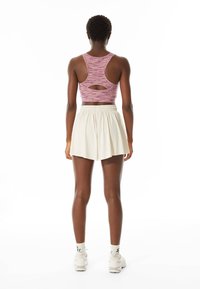 Personne athlétique debout de dos, portant un crop top rose à dos nageur, un short plissé crème, des chaussettes blanches et des baskets blanches.