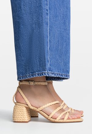 Fod iført beige flettet blokhæls sandal med ankelrem, kombineret med blå denimjeans, croppet over anklen.