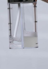 Conteneur de mélange en plastique transparent avec un couvercle en métal, comportant des graduations. Un côté montre un liquide bouillonnant, l'autre est vide.