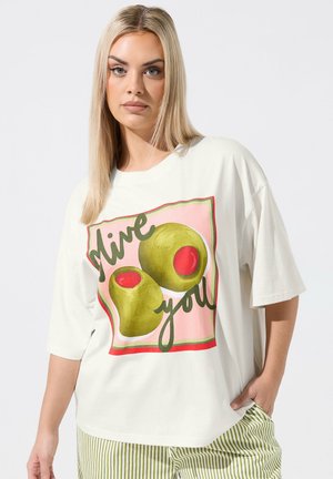 Femme blonde portant un t-shirt blanc oversize décoré d'olives vertes et du texte « olive you », assorti à un pantalon rayé vert et blanc.