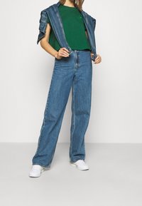 T-shirt vert à manches courtes avec un design de smiley, associé à un jean large bleu clair et des baskets blanches, le tout surplombé d'une veste en denim.