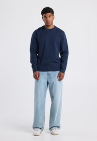 Meripihkala crewneck-sweater, rento muotoilu, yhdistetty vaaleansinisiin leveälahkeisiin farkkuihin ja valkoisiin tennareihin, seisoo neutraalia taustaa vasten.