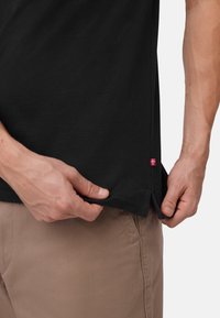Zwarte katoenen t-shirt met zijsplitten en een klein rood-wit logo-label aan de zoom, vastgehouden door een hand nabij beige broeken.