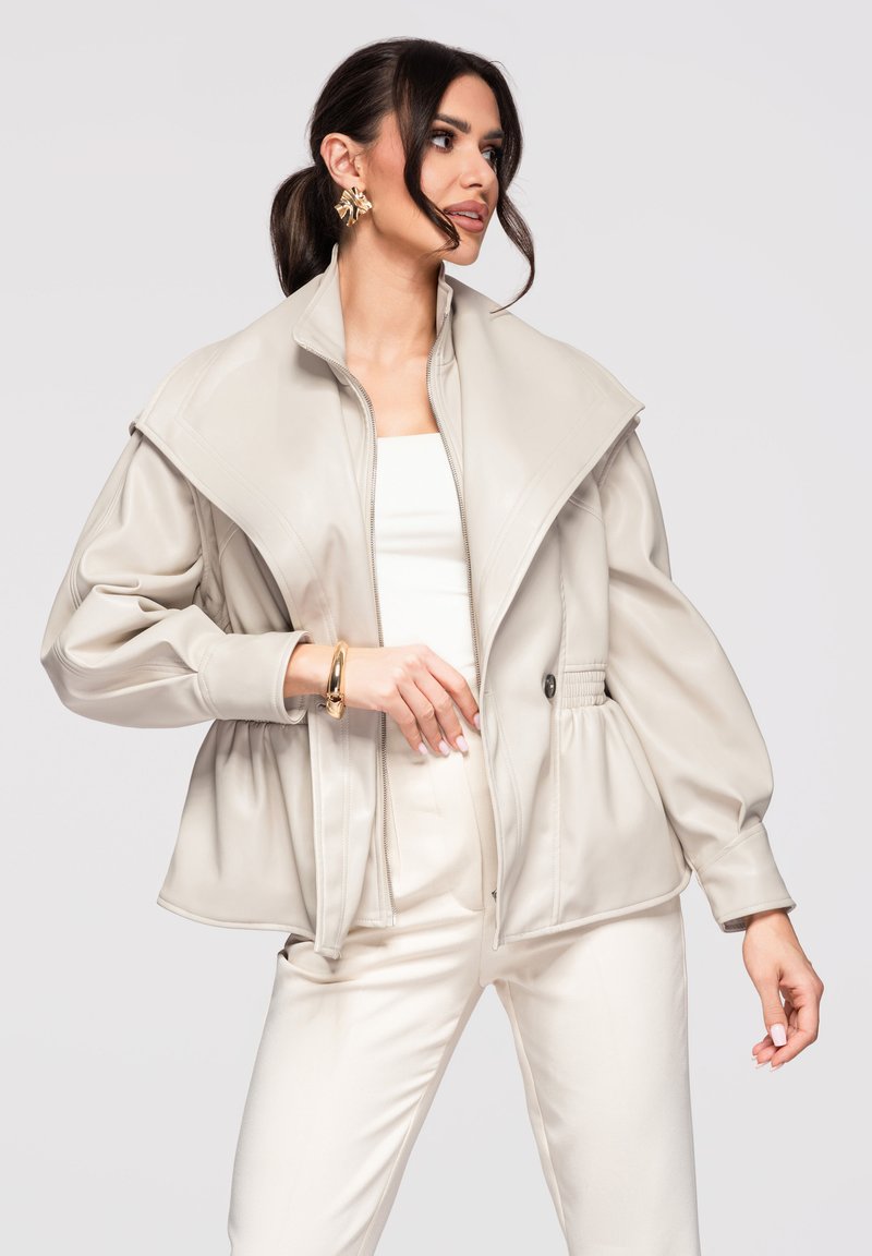 Mujer con chaqueta beige de gran tamaño y cuello ancho, camiseta blanca, pantalones a juego, pendientes de flor dorada y pulsera, posando frente a un fondo liso.