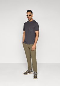 Tricou scurt de culoare gri închis cu buzunar pe piept, pantaloni verzi măslinii și pantofi de drumeție negri cu accente bej, asortați cu ochelari de soare.