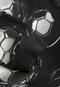 Čierna atletická tkanina s lesklým vzorom futbalových loptičiek. Rukáv je rebrovaný, čím poskytuje textúrny kontrast k hladkému materiálu.