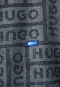 Tela de la marca Hugo con un fondo gris oscuro y un logotipo de texto en gris claro repetido. Pequeña etiqueta tejida azul con letras blancas.