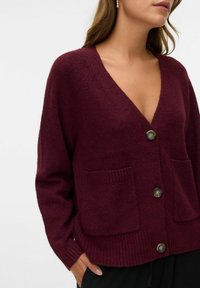 Burgundy pleteni kardigan ima duboki V-izrez, velike prednje džepove i tri gumba s teksturiranim završetkom. Mekani, udoban materijal.