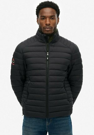 Man die naar voren kijkt, draagt een zwart doorgestikt donsjack met een hoge kraag en een blauwe spijkerbroek, tegen een effen achtergrond.