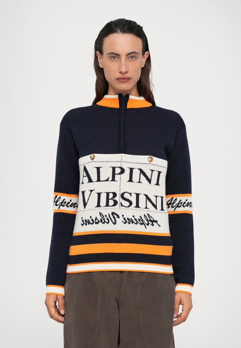 Ženska v temnonavježno-modri, oranžni in beli zip-up puloverju s napisom "Alpini Vibsini" in rjavih hlačah, stoji pred enostavno belo ozadje.