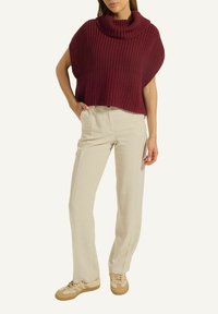 Frau trägt einen ärmellosen, kastanienbraunen, grob gestrickten Rollkragenpullover, beiger, hochtaillierter, gerader Hose und beigen Sneakern.