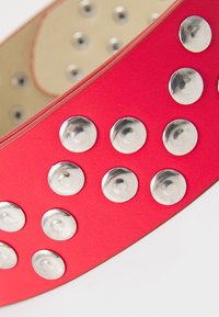 Cintura sintetica rossa con borchie circolari argentate lungo tutta la sua lunghezza, con una texture liscia e un interno beige a contrasto.