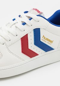 Basket blanche avec des bandes chevrons rouges et bleues, lacets blancs, doublure intérieure bleue et logo doré "hummel Handball Perfekt" sur le côté.