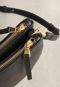 Sac bandoulière en cuir noir avec deux fermetures éclair dorées et une boucle dorée reliant la bandoulière réglable au corps du sac.