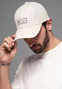 Witte honkbalpet met een gestikte logolabel aan de voorkant. Zacht materiaal, gebogen klep en zes ventilatiegaten bovenop.