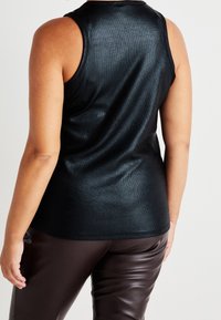 Sort ærmeløs top med en glansfuld, tekstureret finish; blødt stof; tætsiddende design. Båret med mørkebrune læderbukser, vist fra bageste synsvinkel.