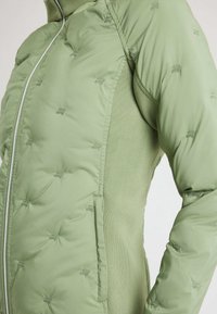 Chaqueta acolchada verde claro con pequeños logotipos acolchados, cremallera central y paneles laterales de punto, mostrada desde la mitad del torso hasta la parte superior del brazo.