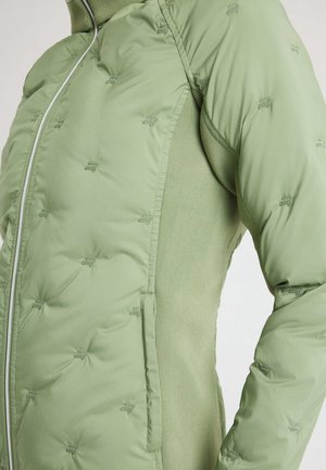 Giacca imbottita verde chiaro con piccoli loghi trapuntati, zip centrale e pannelli laterali in maglia, mostrata dalla parte centrale del torace fino al braccio superiore.
