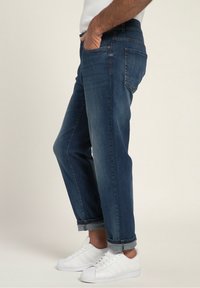 Hombre con jeans oscuros doblados y zapatillas blancas, de pie con una mano en el bolsillo, frente a un fondo sencillo.