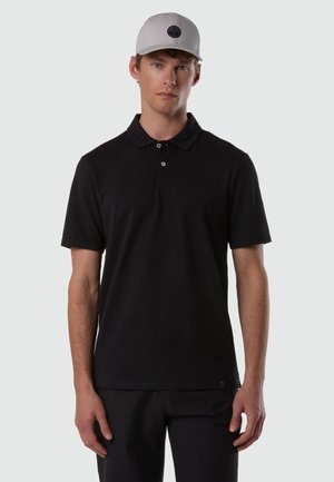 Junger Mann, der nach vorne blickt, trägt ein schwarzes kurzärmeliges Poloshirt und eine hellgraue Baseballkappe vor einem schlichten Hintergrund.