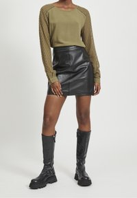 Blouse vert olive avec manches texturées, mini-jupe en cuir noir, et bottes noires montantes à fermeture éclair et semelles en caoutchouc.