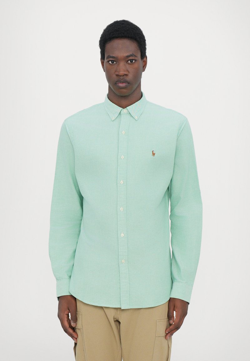 Chemise boutonner vert clair avec un col, des manches longues et un petit logo brodé sur la poitrine gauche ; présentée sur un mannequin.