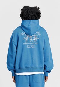 Modrý pulover s kapucí a přední kapsou, s bílým grafickým textem a designem na zádech. Bavlněný materiál s žebrovanými manžetami a lemováním.