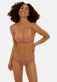 Soutien-gorge en dentelle rose blush avec culotte assortie. Le soutien-gorge présente des bonnets rembourrés, des bretelles réglables et une broderie florale ; la culotte est en tissu transparent avec des coutures décoratives.