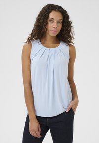 Gaiši zila bez piedurknēm tops ar pleitu prieksdaļu, mīksts audums, apaļa kakla izgriezums un brīvs piegriezums, apvienots ar tumšiem džinsiem.