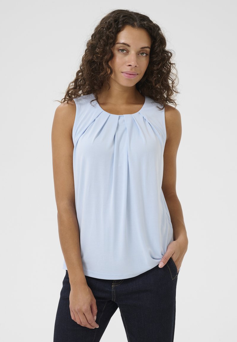 Gaiši zila bez piedurknēm tops ar pleitu prieksdaļu, mīksts audums, apaļa kakla izgriezums un brīvs piegriezums, apvienots ar tumšiem džinsiem.