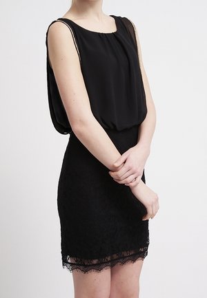 Femme portant une robe noire sans manches avec un haut drapé et une jupe bordée de dentelle, les bras croisés légèrement devant elle.