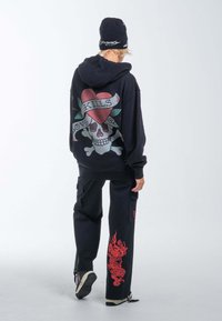 Felpa nera con grafica di teschio, cuore e accenti floreali; abbinata a pantaloni cargo neri con un design di drago rosso. Includendo un berretto.