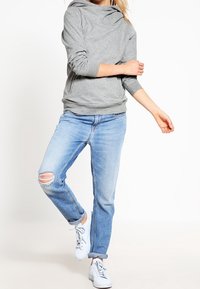 Sweat à capuche gris avec un col cheminée, manches longues et poche kangourou. Associé à un jean bleu clair déchiré et des baskets blanches.