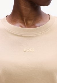 Beige pamut póló bordázott környakkal és texturált "BOSS" logóval a mellkason. Puha, sima tapintású anyag.