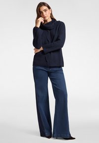 Frau steht mit der Hand am Kinn, trägt einen marineblauen, gerippten Pullover mit Knopfausschnitt und weit geschnittene, dunkelblaue Jeans mit schwarzen Absätzen.