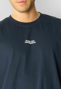 Mørkeblå bomull T-skjorte med hvit trykt logo "CLEAN CUT COPENHAGEN" på brystet. Rund hals og avslappet passform.