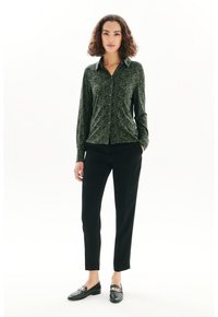 Chemise à motifs verts avec col, associée à un pantalon noir. Des mocassins noirs avec une chaîne dorée complètent la tenue.