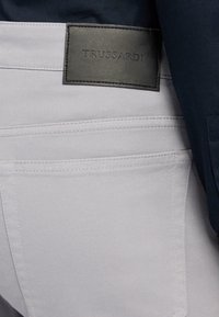 Jeans in denim grigio con una texture liscia, caratterizzati da un prominente etichetta in pelle nera con la scritta "TRUSSARDI" sul retro.