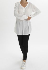 Blouse blanche à manches longues avec un col en V et des détails de boutons, associée à un pantalon noir ajusté et des baskets blanches, créant un look superposé.